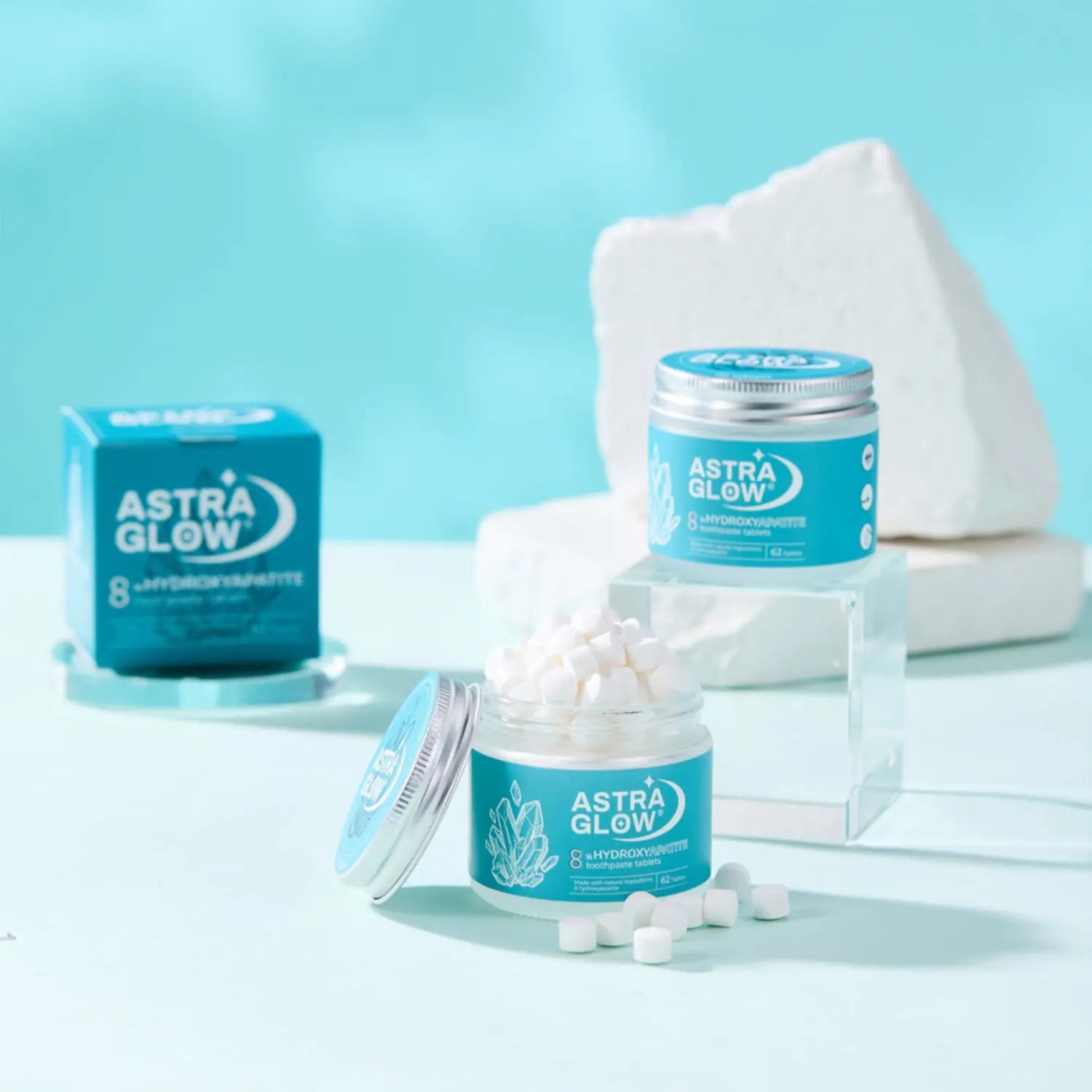 AstraGlow 8% hydroxyapatite toothpaste tablets in teal jars with white tablets scattered, set against a clean blue background.